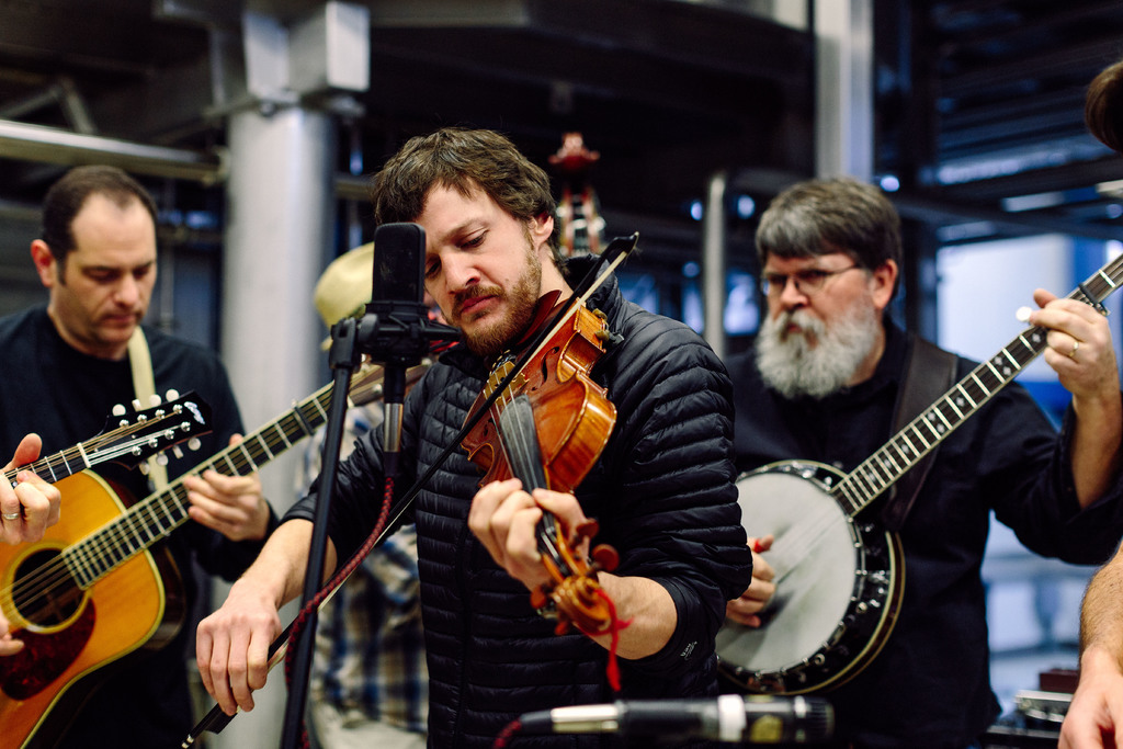 Eine Gruppe von Musikern in schwarzer Kleidung, die verschiedene Instrumente spielen, mit einem Geigenvirtuosen vor einem Mikrofon und einem Gebäude im Hintergrund.