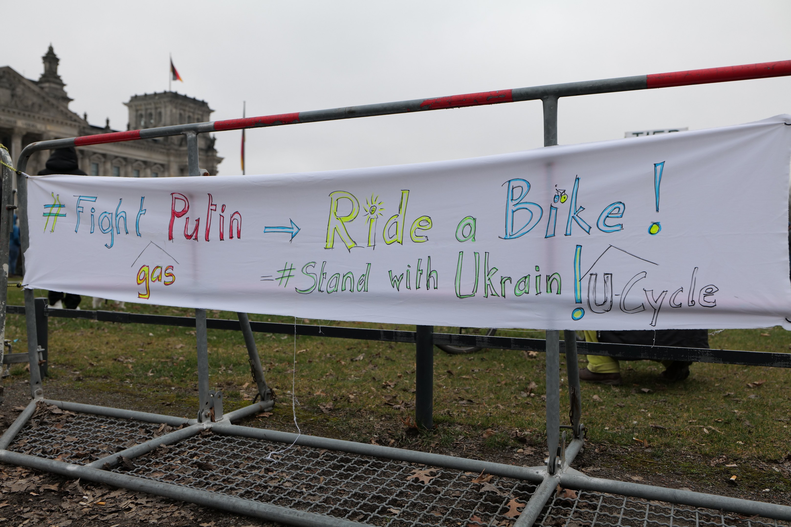 Gruppe von Menschen mit einem Banner "Kämpfe gegen Putin, fahre Rad, stehe mit ukrainischem U-Cycle" vor einem Metallzaun, trockenen Blättern, einem Gebäude mit Fenstern, einem Fahnenmast mit einer Flagge und einem bewölkten Himmel.
