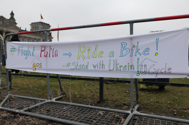 Gruppe von Menschen mit einem Banner "Kämpfe gegen Putin, fahre Rad, stehe mit ukrainischem U-Cycle" vor einem Metallzaun, trockenen Blättern, einem Gebäude mit Fenstern, einem Fahnenmast mit einer Flagge und einem bewölkten Himmel.