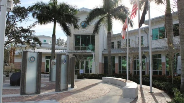 Eingang zu einem Gebäude mit Palmen, Pfählen, Flaggen und einem Logo, wahrscheinlich das Florida Department of Public Safety, vor einem Himmel-Hintergrund.