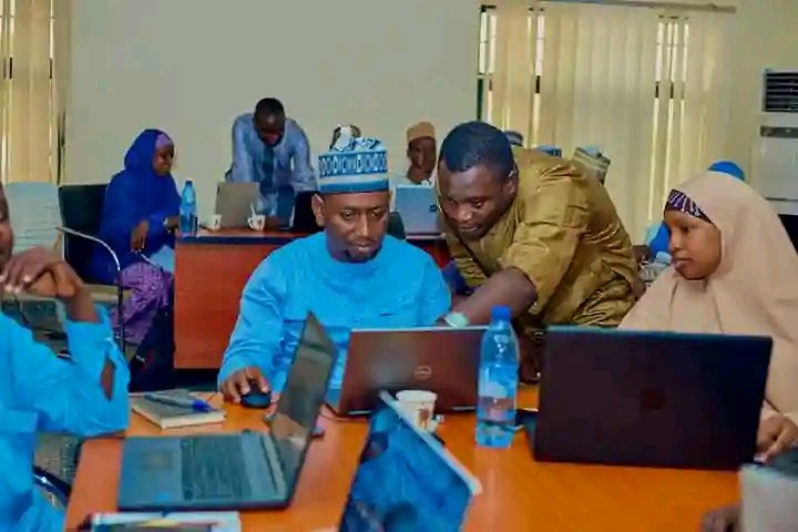 Gruppe von Menschen, die um einen Tisch mit Laptops und Bürobedarf sitzen, arbeiten in einem Raum mit Fenstern, Vorhängen und einer Klimaanlage.