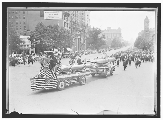 Schwarzes und weißes Foto von einer Parade mit einem Löschfahrzeug in der Mitte, umgeben von Menschen mit Fahnen, Bäumen, Gebäuden und einer Tafel im Hintergrund.