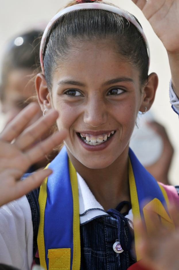 Ein junges Mädchen in Schuluniform, das fröhlich winkt und im Hintergrund etwas unscharfe Gestalten zu sehen sind.