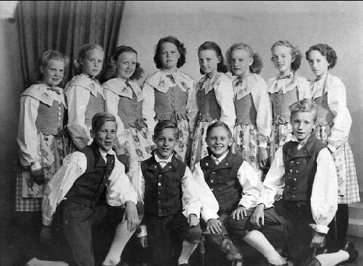 Schwarz-weißes Foto einer Gruppe von Kindern, die vor einer Vorhangwand posieren, mit dem Text "1930er-Kinder-Schule" unten.
