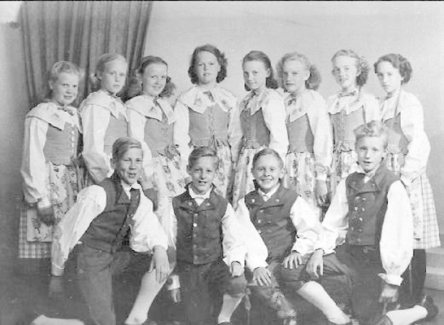 Schwarz-weißes Foto einer Gruppe von Kindern, die vor einer Vorhangwand posieren, mit dem Text "1930er-Kinder-Schule" unten.