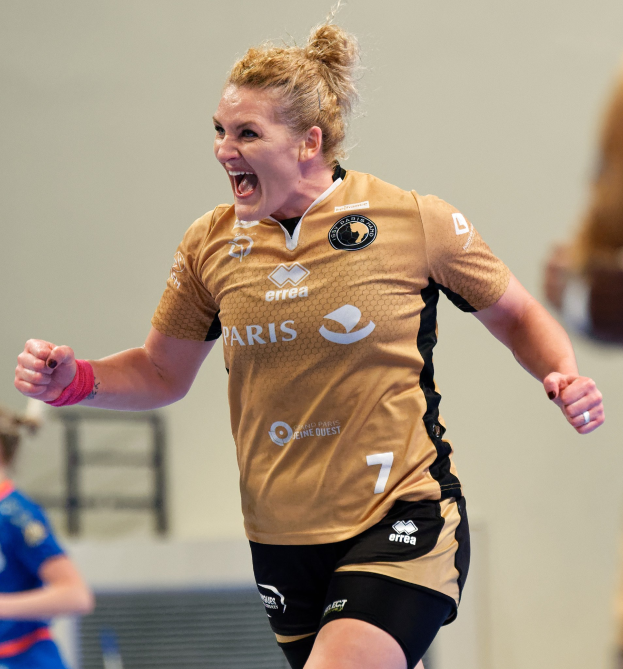 Eine Frau in braunem T-Shirt und schwarzen Shorts rennt und feiert auf einem Futsalplatz mit zwei anderen Frauen im Hintergrund.
