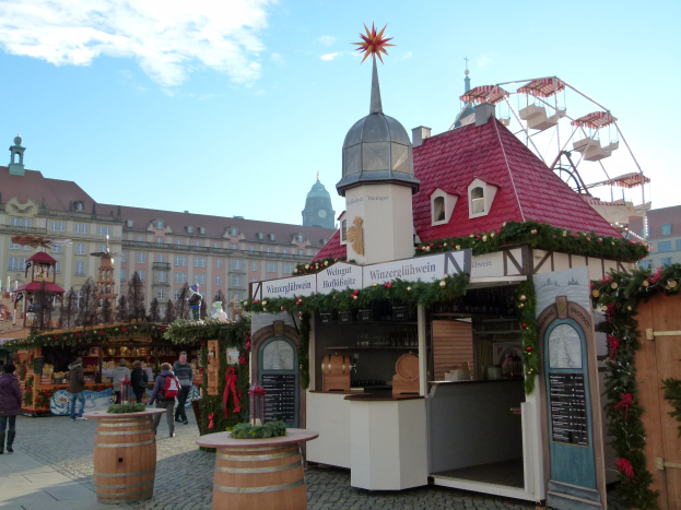 Ein geschäftiger Weihnachtsmarkt in Nürnberg, Deutschland, mit Menschen um dekorierte Stände, festliche Lichter und Schmuck, mit einem Riesenrad, Gebäuden mit Fenstern und einem bewölkten Himmel.