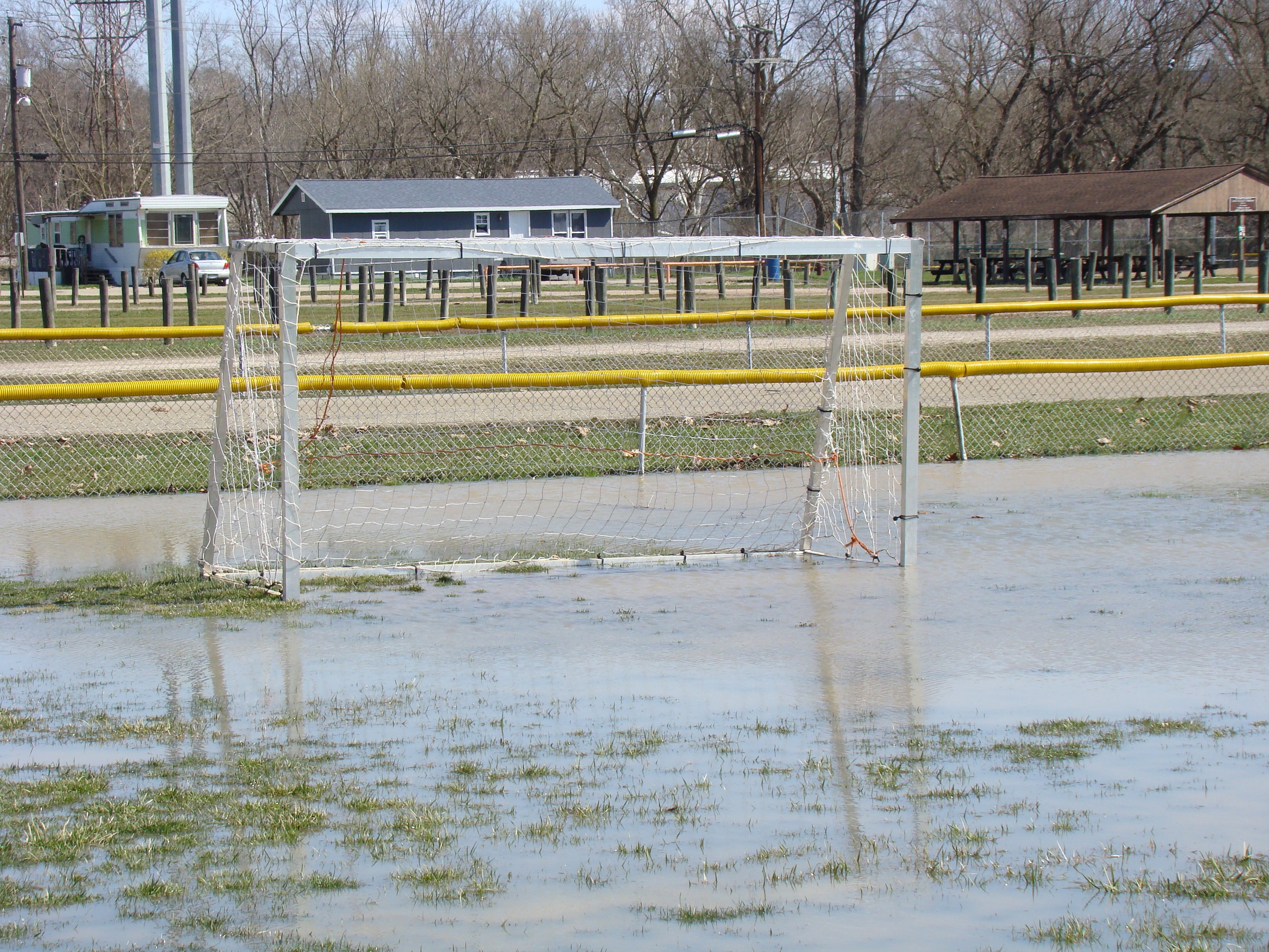 Ein Fußballtor steht in der Mitte eines überfluteten Feldes, umgeben von Gras und Wasser, vor einer Kulisse aus Hütten, Pfählen, Bäumen, Fahrzeugen und einem klaren blauen Himmel.