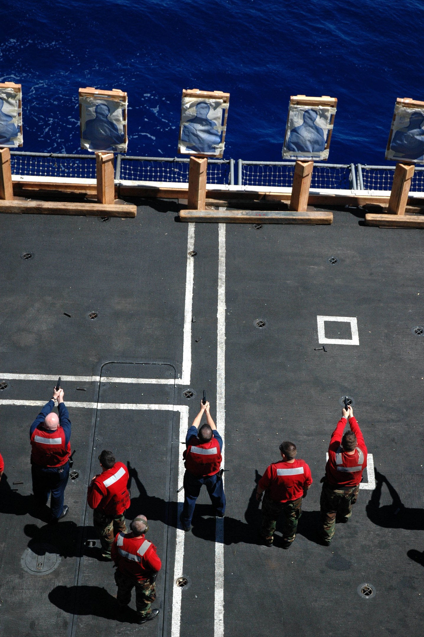 Männer in roten Jacken mit Gewehren auf einem Flugzeugträger mit einem Ozean und einem Zaun im Hintergrund.