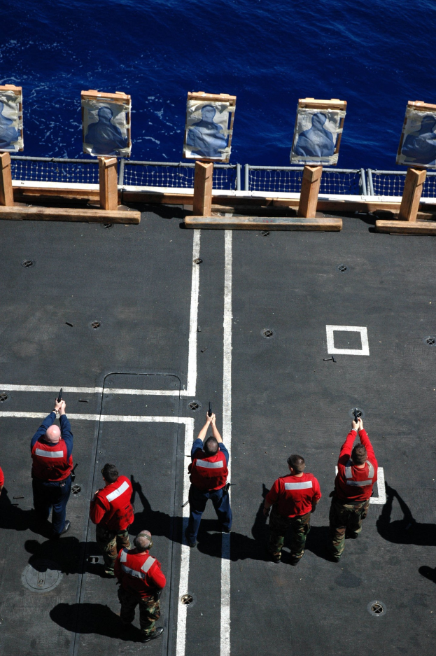 Männer in roten Jacken mit Gewehren auf einem Flugzeugträger mit einem Ozean und einem Zaun im Hintergrund.