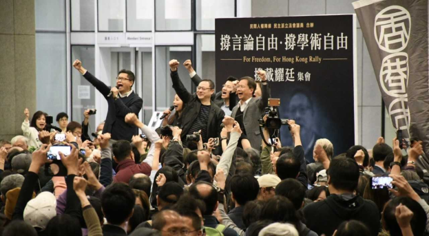 Eine Gruppe von Menschen steht vor einer Menge mit Kameras, Handys und Mikrofonen, mit einem Banner, einer Wand und Glastüren im Hintergrund, wahrscheinlich protestieren sie gegen das Verbot des Hongkonger Auslieferungsgesetzes.