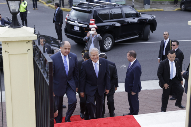 Gruppe von formell gekleideten Männern, die eilig über einen roten Teppich gehen, mit einem Fotografen im Vordergrund, Stadtstraßenszene im Hintergrund.