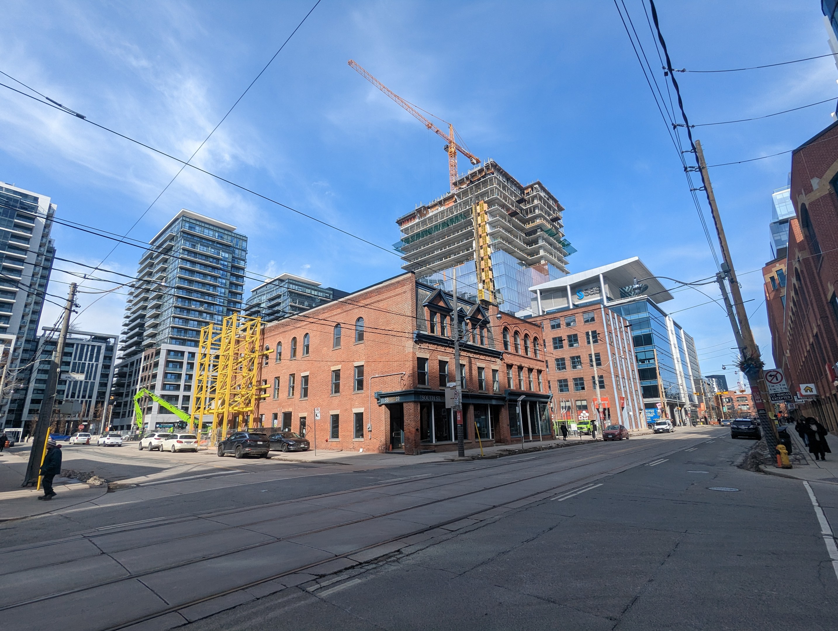 Eine belebte Stadtstraße mit fahrenden Fahrzeugen, Fußgängern auf dem Gehweg, Strommasten mit Drähten, Schildern und Gebäuden, mit einem Baukran im Hintergrund bei bewölktem Himmel in Toronto.