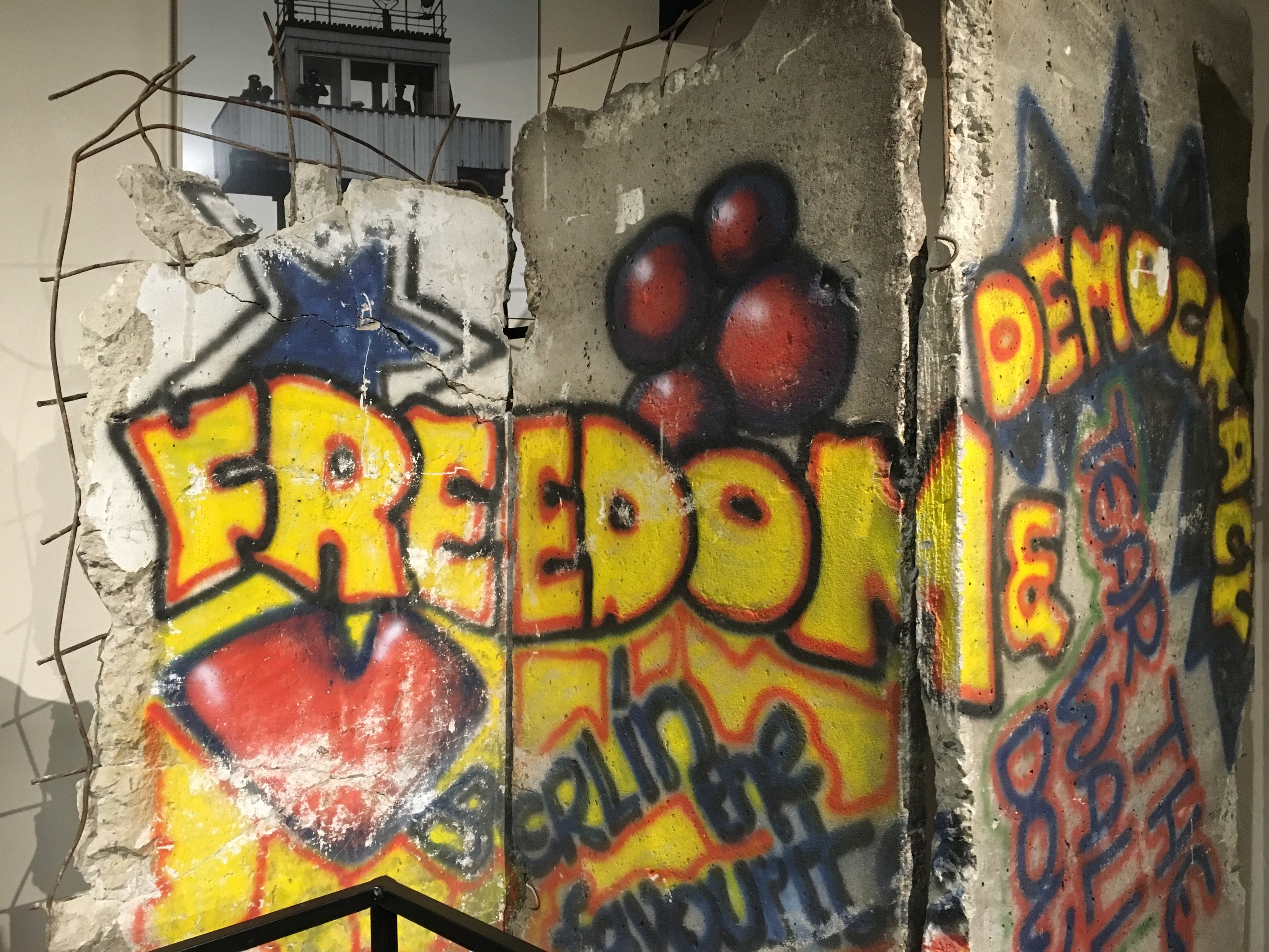Mauer mit Graffiti in fetten schwarzen Buchstaben "Freiheit und Demokratie", eine Reling unten und ein Gebäude mit Fenstern im Hintergrund.