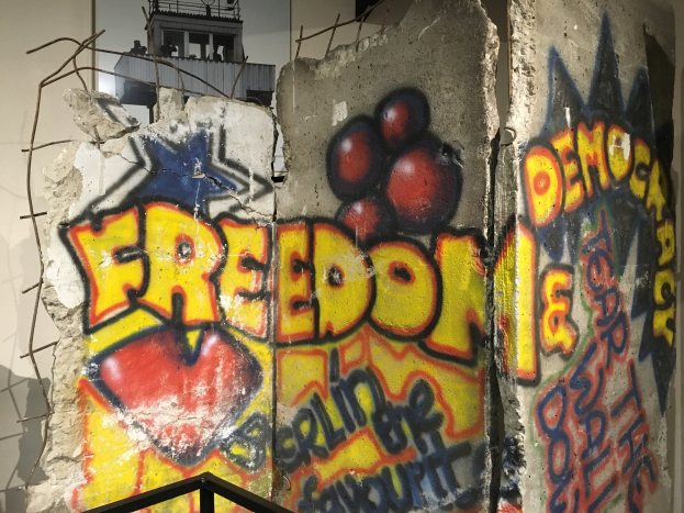 Mauer mit Graffiti in fetten schwarzen Buchstaben "Freiheit und Demokratie", eine Reling unten und ein Gebäude mit Fenstern im Hintergrund.