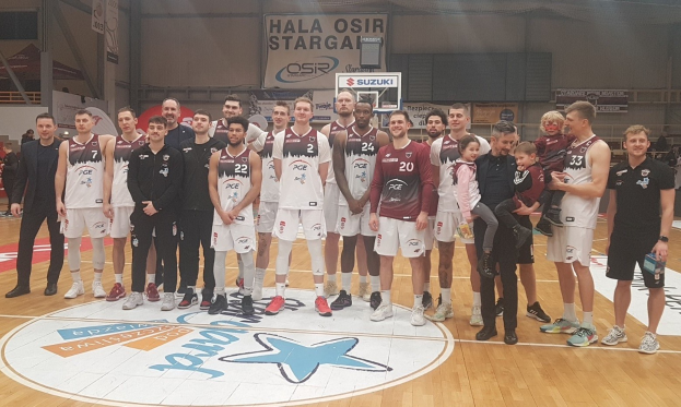 Gruppe von Männern und Kindern auf einem Basketballfeld mit Schildern "Hala Osir Starga", sitzende Zuschauer und Basketballausrüstung im Hintergrund.