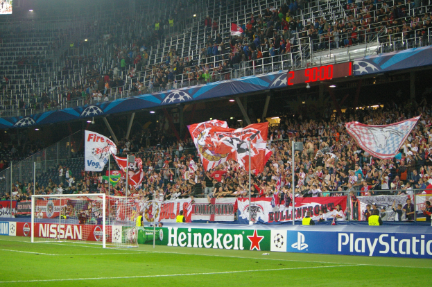Ein Fußballspiel in einem hell erleuchteten Stadion mit einer großen Zuschauermenge, einem Tor in der Mitte, Bannern drumherum und einem Bildschirm oben links.