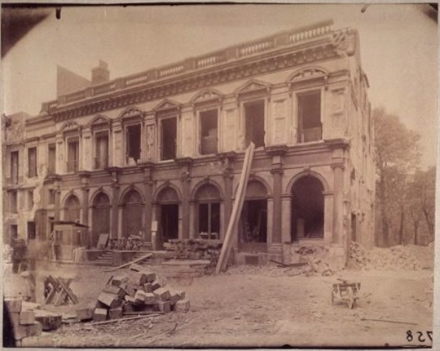 Schwarzes und weißes Foto eines abbruchreifen Gebäudes mit Fenstern, Säulen, Bögen, hölzernen Gegenständen, einem Karren, Bäumen und Himmel mit Text am unteren Rand.