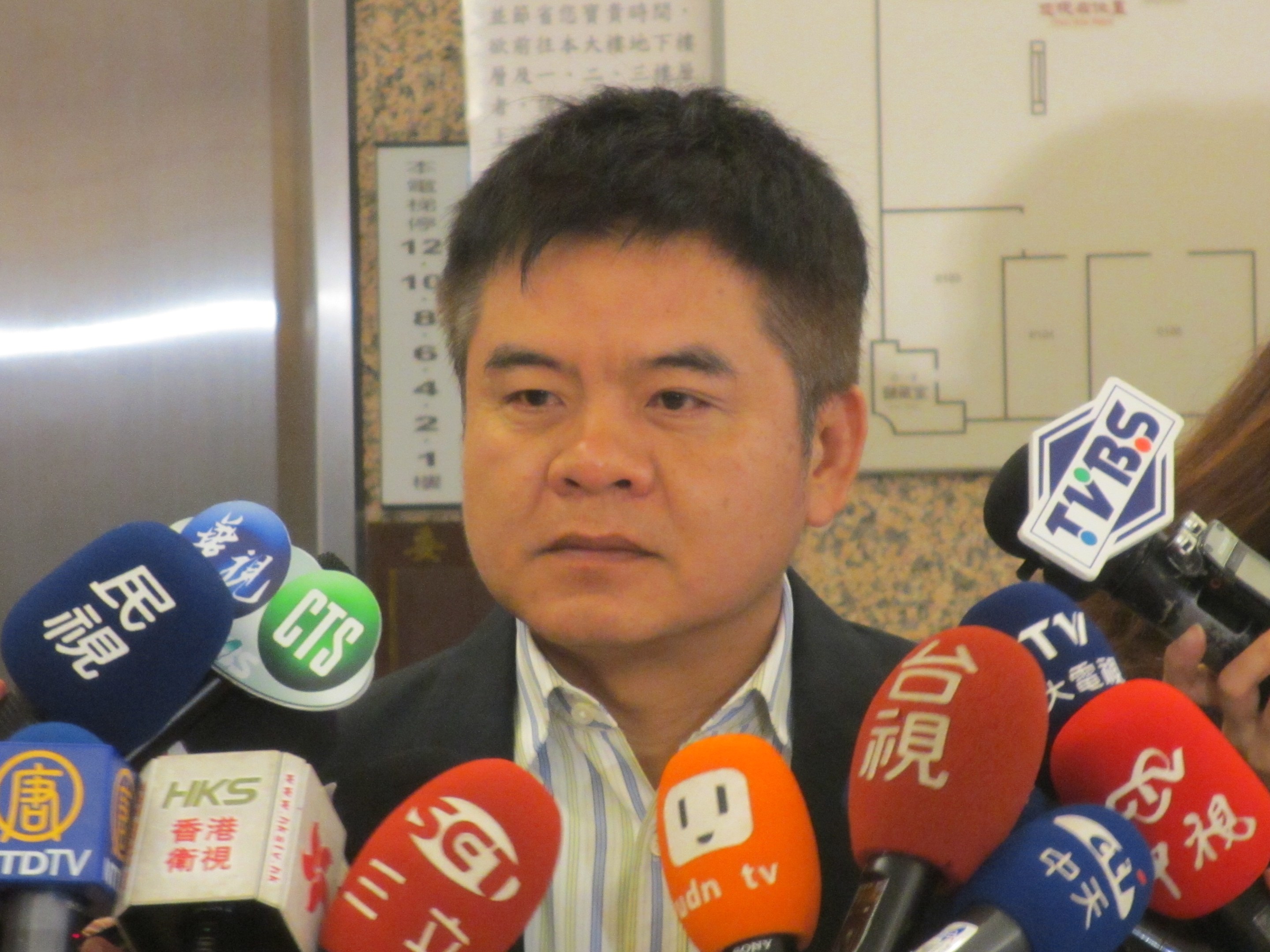 Ein Mann im Anzug und Krawatte spricht vor einer Wand mit einer Tafel zu Reportern, mit einer Frau mit einer Kamera rechts daneben.