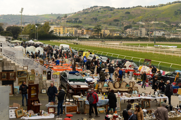 Eine große Menschenmenge auf einem Outdoor-Flohmarkt mit Tischen, auf denen Gegenstände wie Foto Rahmen und Stühle ausgelegt sind, umgeben von geparkten Fahrzeugen, Geländern, Treppen, Bäumen, Gebäuden, Laternenmasten, Hügeln und einem bewölkten Himmel.
