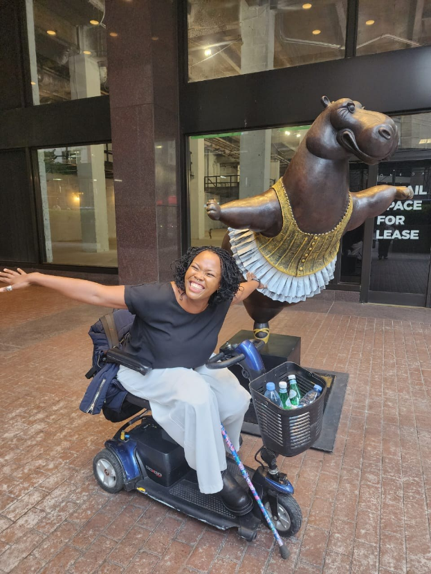 Eine Frau in einem Mobilitätsscooter mit ausgestreckten Armen vor einer Nilpferd-Statue, die ein schwarzes T-Shirt und weiße Hosen trägt, mit einem Korb voller Flaschen neben ihr, vor Glastüren mit Deckenlampen.