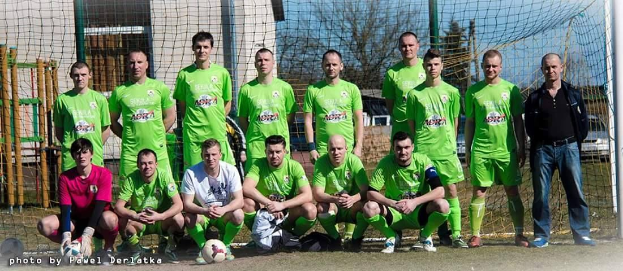 Eine Gruppe von Männern in grünen T-Shirts steht auf einem Fußballfeld in der Nähe eines Torpfostens, einige hocken, mit einem Ball davor; der Hintergrund enthält Bäume, Gebäude und Fahrzeuge unter dem Himmel.