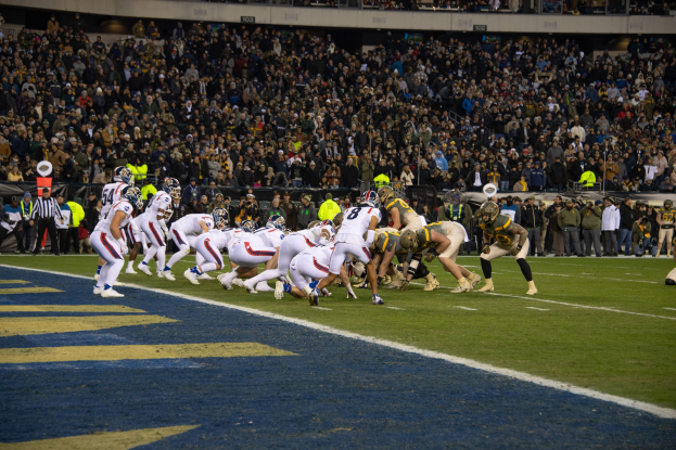 Fußballspieler in Sportkleidung und Helmen auf dem Feld während eines Spiels in einem Stadion mit Zuschauern und Tafeln im Hintergrund.