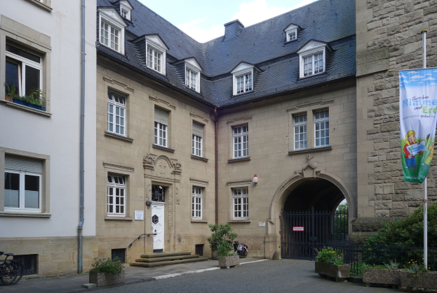 Hof mit einem Gebäude mit Fenstern, einer Tür, einer Treppe, einem Metallzaun, einer Fahne auf einem Pfahl, Topfpflanzen, parkenden Fahrrädern, Bäumen und einem bewölkten Himmel.
