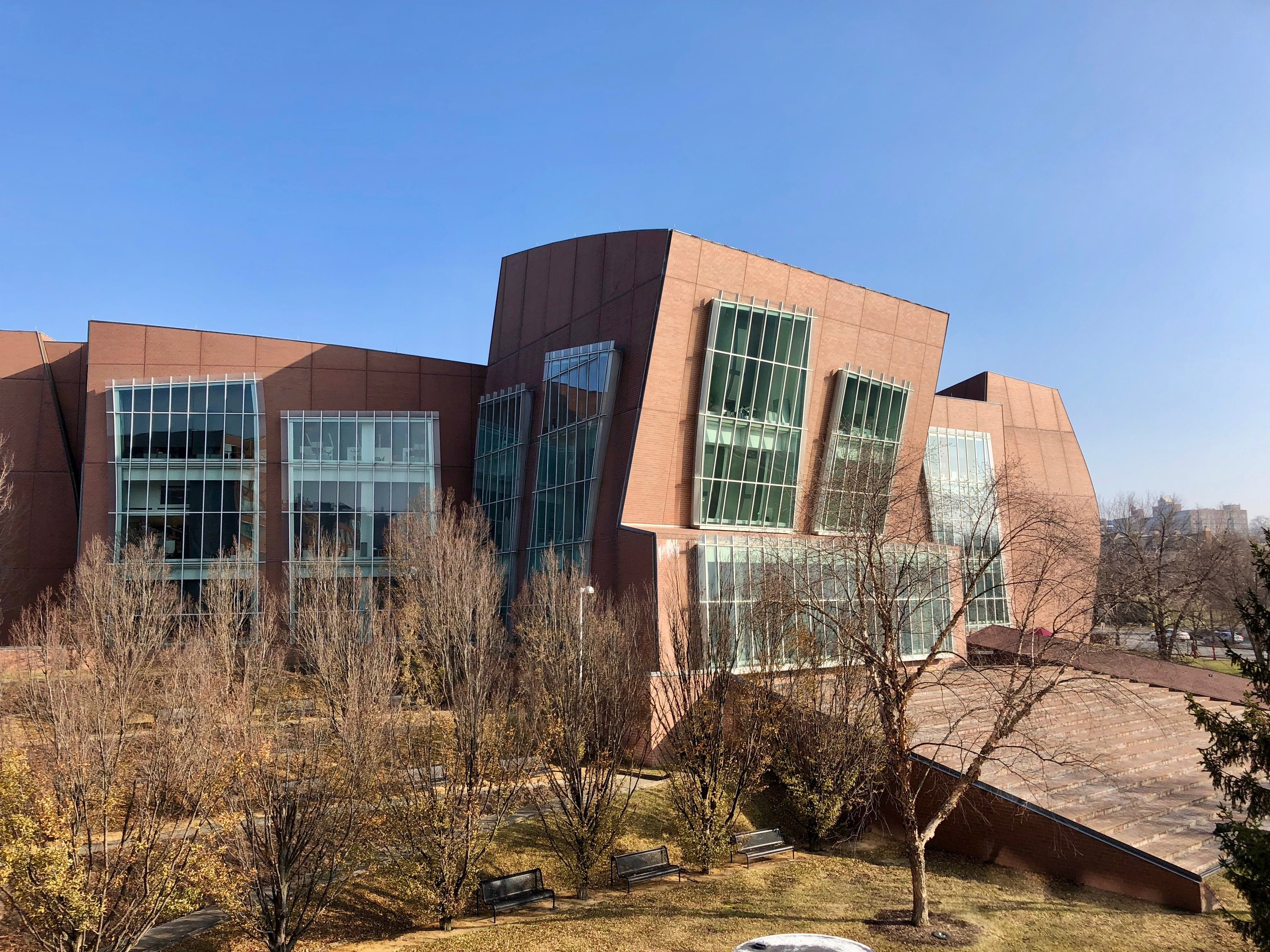 Großes mehrstöckiges Universitätsgebäude mit zahlreichen Fenstern, umgeben von Bäumen, Bänken, Gras und einem Lichtmast, mit anderen Gebäuden und einem klaren blauen Himmel im Hintergrund.