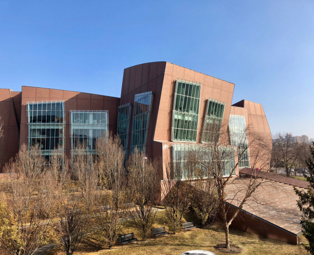 Großes mehrstöckiges Universitätsgebäude mit zahlreichen Fenstern, umgeben von Bäumen, Bänken, Gras und einem Lichtmast, mit anderen Gebäuden und einem klaren blauen Himmel im Hintergrund.