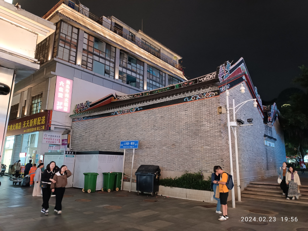 Menschen gehen nachts eine Straße entlang, beleuchtet von Straßenlaternen und umgeben von Gebäuden, Schildern, Mülltonnen, Pflanzen, Bäumen und einem bewölkten Himmel, einige tragen Taschen.