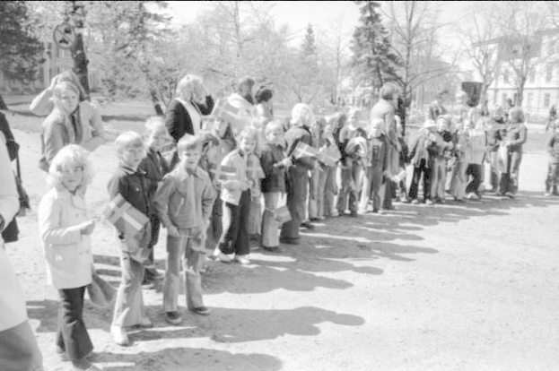 Schwarz-weißes Bild einer Gruppe von Menschen, die Fahnen auf einer Schotterstraße während einer Protestmarsch an einer Schule halten, mit Bäumen, Gebäuden und einem klaren Himmel im Hintergrund.