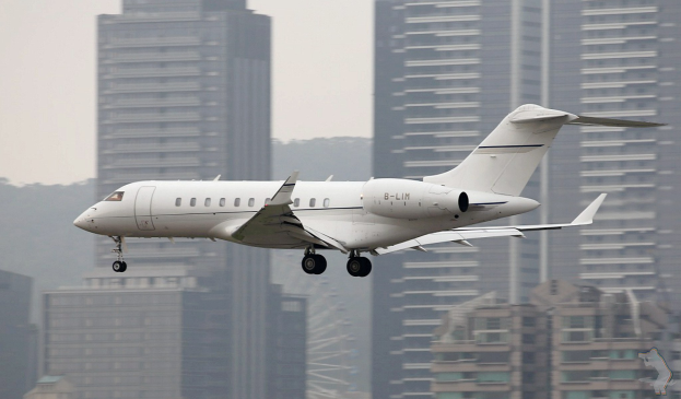 Wei├čer Privatjet über einer Stadt mit hohen Geb├Ąuden im Hintergrund, Himmel oben sichtbar, Wasserzeichen in der rechten unteren Ecke.