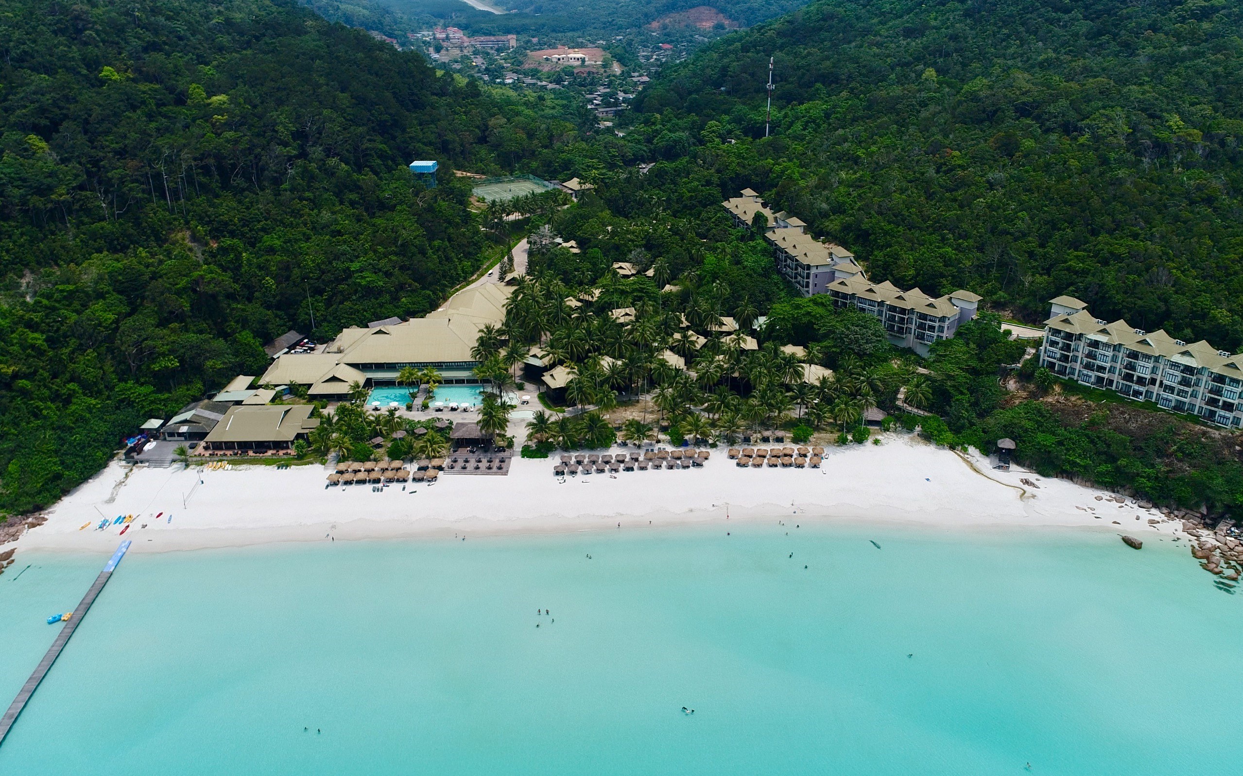 Luftaufnahme eines Strandresorts in Koh Samui, Thailand, mit einer Brücke auf der linken Seite, Gebäuden, Bäumen, Hügeln und ein paar Menschen am Strand.