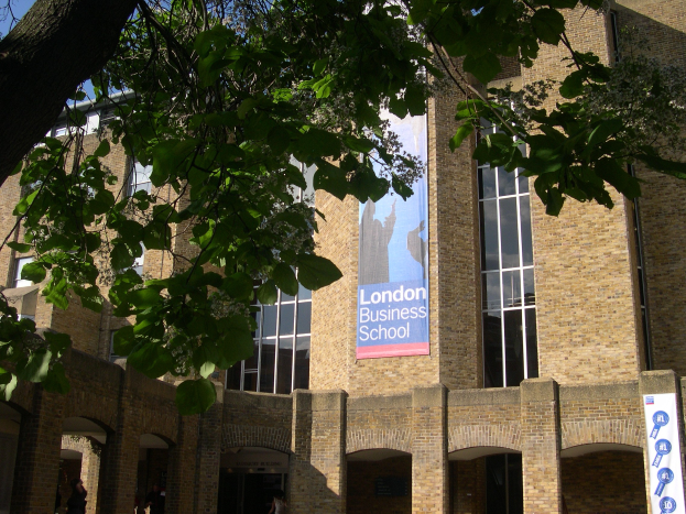 Großes Backsteingebäude mit "London Business School"-Schriftzug, umgeben von Bäumen unter einem klaren blauen Himmel.