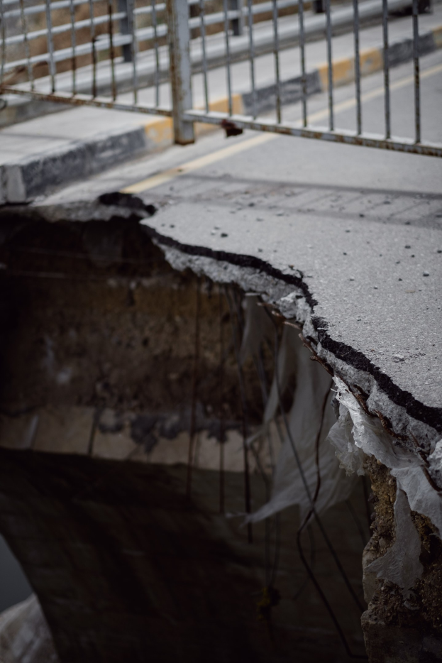 Großer Riss an der Seite einer Brücke mit Metallgeländer und einer Straße im Hintergrund.