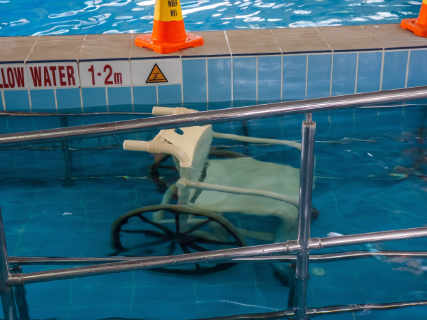 Schwimmbad mit einem Stuhl in der Mitte, umgeben von Sicherheitskegeln und einem Geländer, mit einem Schild an der Wand, das über bereitgestellte Schwimmausrüstung informiert.