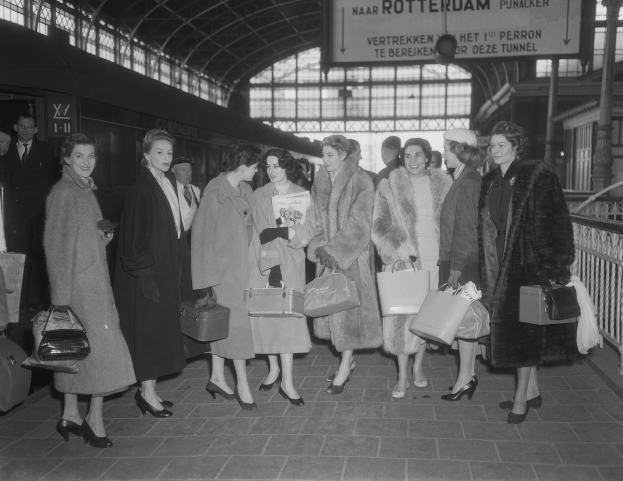 Schwarz-weißes Bild von Frauen an einem Bahnhof mit Taschen, einem Zug auf der linken Seite und Geländern und einer Tafel auf der rechten Seite.