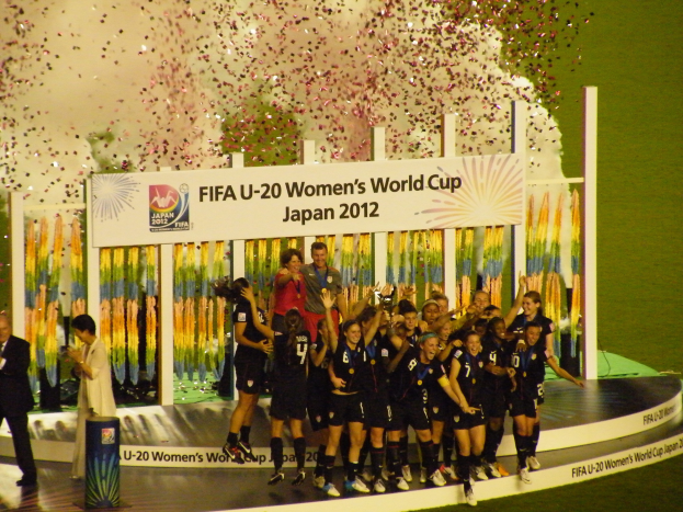 Gruppe von Frauenfußballspielerinnen in schwarzer Uniform auf einer Bühne stehend, die mit einer Menge, die Kameras hält, feiern.