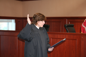 Eine Frau in schwarzer Robe steht vor einem Richtertisch und hält eine Akte in der Hand, mit einem Mikrofon und einer Flagge auf dem Tisch, Stühle und ein Fenster dahinter.