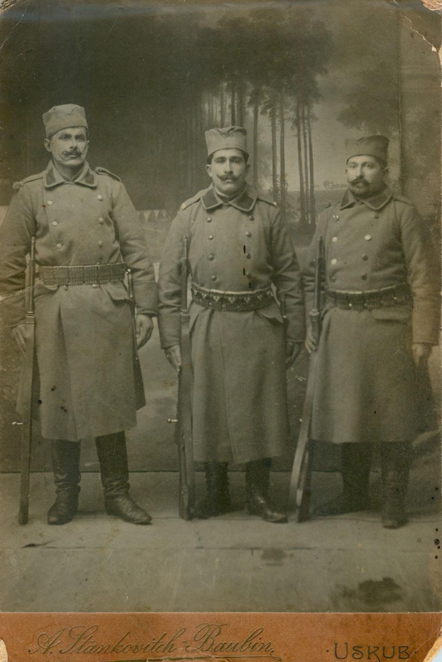 Drei Männer in Militäruniformen stehen nebeneinander, jeder trägt eine Mütze und hält ein Gewehr, mit Bäumen im Hintergrund und dem Text "Sergei Stankovitch Baubin und seine Söhne" unten.