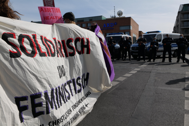 Eine Gruppe von Menschen marschiert auf einer Straße, hält ein Banner mit der Aufschrift "Solidarität und Feminismus", mit parkenden Fahrzeugen und Gebäuden im Hintergrund unter einem klaren blauen Himmel.