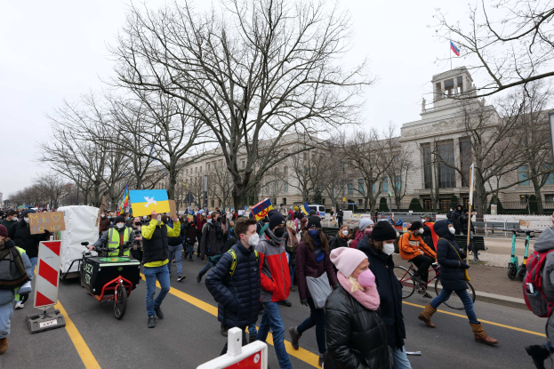 Eine große Protestdemonstration mit Menschen, die eine Straße in Washington, D.C. entlangmarschieren, einige halten Schilder und fahren Fahrräder, mit Bäumen und einem klaren blauen Himmel im Hintergrund.