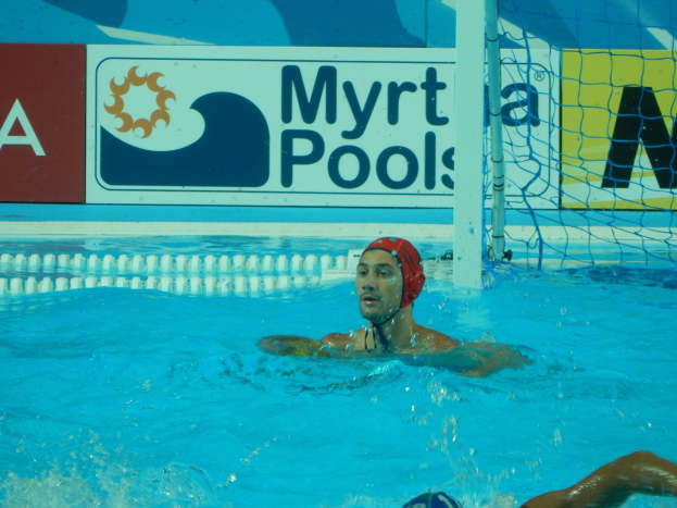 Wasserballspieler in Bewegung mit einer Mütze tragend, der in einem Pool schwimmt, mit einem Netz auf der rechten Seite und einer Werbetafel im Hintergrund.
