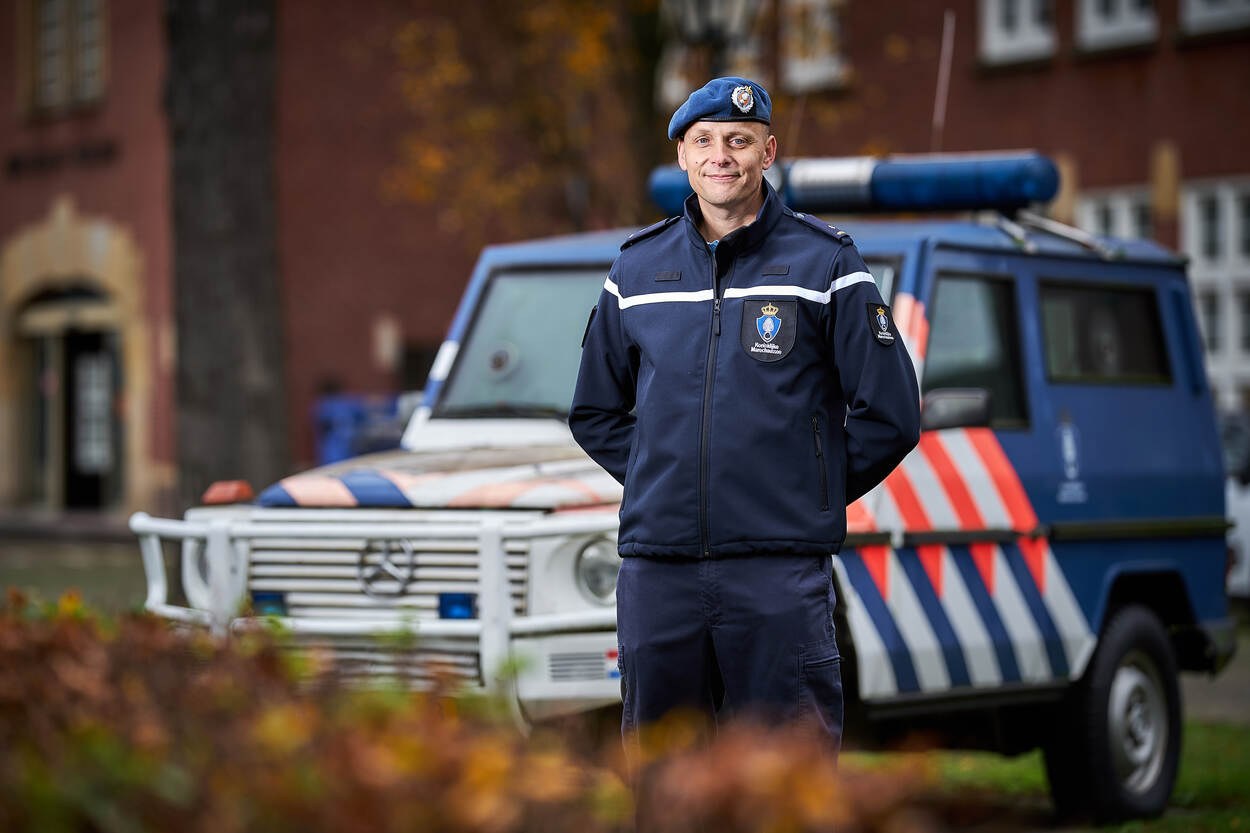Ein Polizeibeamter in Uniform und Mütze steht vor einem Polizeiwagen mit leicht unscharfem Hintergrund und Pflanzen unten.