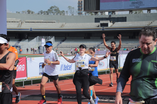 Gruppe von Läufern auf einer Stadionbahn mit Bannern, einem Display, einem Gebäude, Bäumen und einem bewölkten Himmel; einige Läufer tragen Turnschuhe und Hüte.