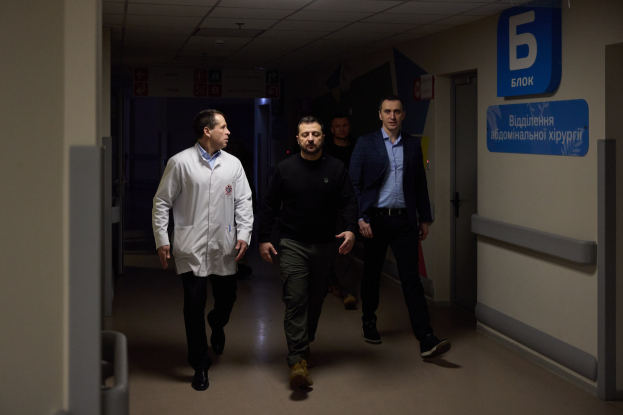 Eine Gruppe von Männern, die einen Krankenhausflur entlanggehen, mit Deckenleuchten, einer Tür an der rechten Wand und Texttafeln an der linken Seite, die besorgt aussehen.