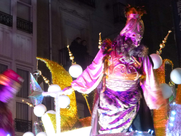 Ein Mann in einem Kostüm steht auf einem geschmückten Wagen bei einem Umzug, beleuchtet von Lichtern, mit einem Stadtgebäude im Hintergrund.