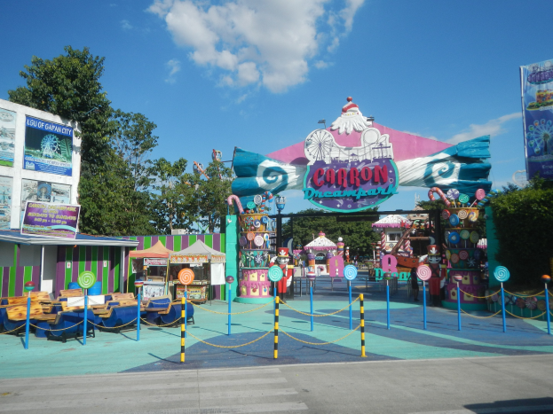 Vergnügungsfahrt am Parkeingang mit Straße, Umzäunung, Buden, Banner, Bäumen und bewölktem Himmel.