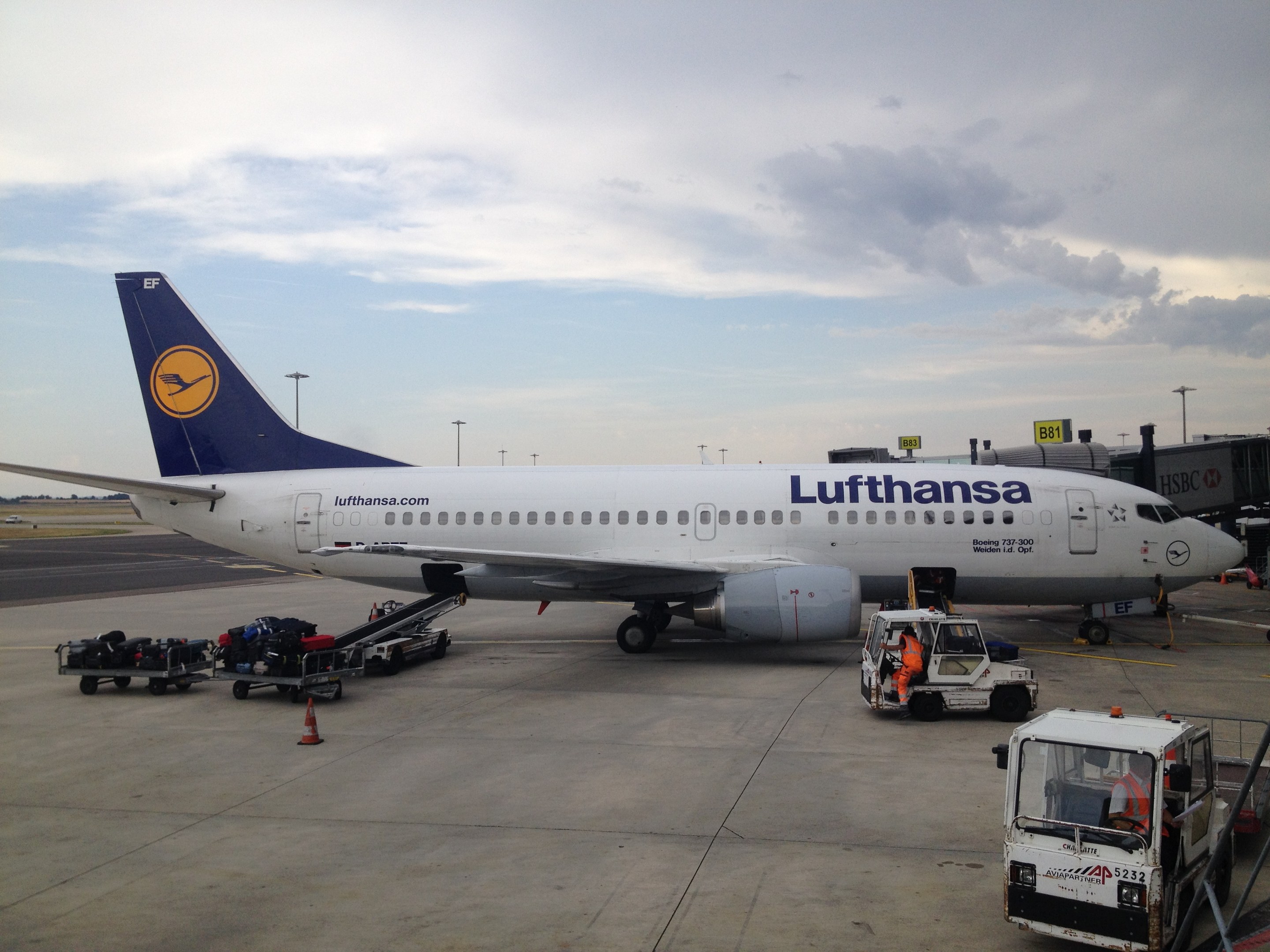 Lufthansa Airbus A320-200 Flugzeug am Frankfurt Airport geparkt mit Bodenfahrzeugen, Verkehrskegeln und Beschilderung im Hintergrund bei bew├Âlktem Himmel.
